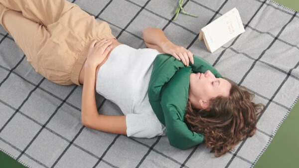 Person lying on a picnic blanket outdoors resting their head on a green infinity pillow while reading a book